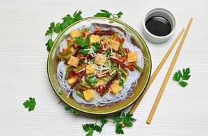 Top View Food Photo of Rice Vermicelli with Tofu, Vegetables and Parsley on Plate next to Cup with Soy Sauce and Chopsticks
