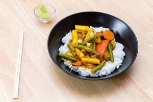 Top View Food Photo of Vegetarian Thai Curry with Rice in Black Ceramic Bowl next to Chopsticks and Small Glass Bowl with Lemon on Wooden Table
