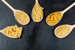Top-view Nahrungsmittel Konzept - unterschiedliche Formen von Pasta in Holzlöffeln auf schwarzem Hintergrund