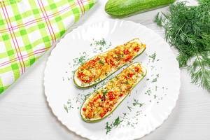 Top view of baked zucchini halves with vegetables, couscous and greens on a white wooden background with fresh zucchini and dill