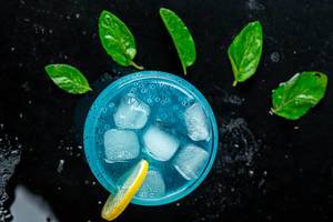 Top view of glass with blue cocktail, ice cubes and mint leaves on black background