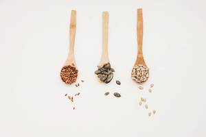 Top view of three wooden spoons with flaxseed, pumpkin seed and sunflower seed on white background