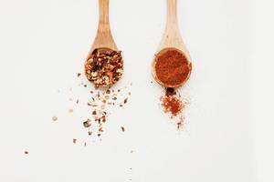 Top view of two wooden spoons with chili flakes and paprika powder on white background