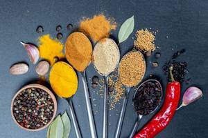 Top view on spices on spoons - curcuma, curry, garlic, mustardseeds and barberries with nutmeg in front on black background