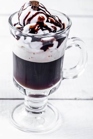 Trinkglas mit einem Sahnekaffee, Schlagsahne, Kaffeebohne und Schokoladensauce auf einem weißen Holztisch