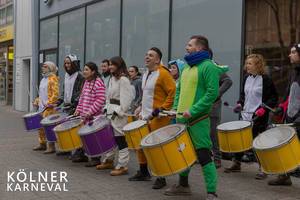Trommelgruppe in Tierkostümen stehen vor einem Geschäft in der Fußgängerzone und machen Musik, neben dem Bildtitel "Kölner Karneval"
