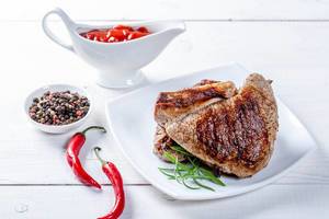 Two hot beef steaks on a white table with chili pepper and sauce