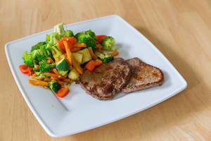 Two mini-steaks with peppers, broccoli, zucchini and carrots on a white plate on a table