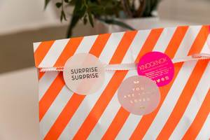 Überraschungstüte als liebevolles Geburtstagsgeschenk in einer weiß-orange-gestreiften Papiertüte mit drei runden Aufklebern