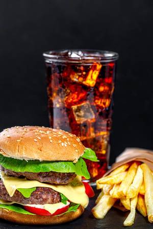 Ungesundes Ernährungskonzept: Hamburger, Cola mit Eiswürfeln und frischen Pommes, vor schwarzem Hintergrund