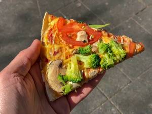 vegan Pizza slice "Kap Verde" from Dominos in Hand with Tomato, broccoli, fresh mushrooms and vegan processed cheese toppings