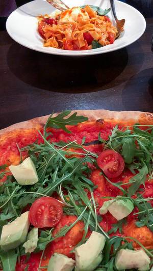 Vegan pizza (without cheese) with avocado, cherry tomatoes, rucola and tomato sauce. In the background, pasta with tomato sauce and cheese