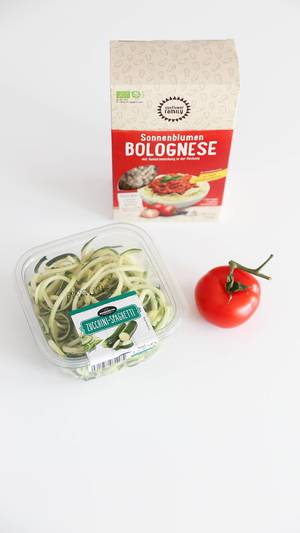 Vegane Bolognese aus Sonnenblumenkernen mit Gewürzmischung, neben einer Tomate und Zucchini-Spaghetti, vor weißem Hintergrund