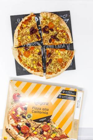 Vegane Pizza mit Bohnen, Mais und Tomaten auf Schieferplatte neben Pizzakarton vor weißem Hintergrund