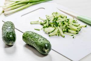 Vegetarisch kochen: ganze und klein geschnittene Gurken auf weißem Schneidebrett mit grünem Messer und ganzen Frühlingszwiebeln