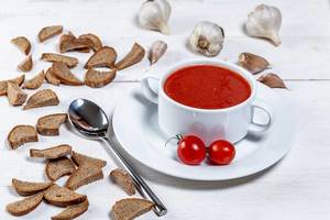 Vegetarische Tomatensuppe in weißer Tasse mit Kirschtomaten, Brotecken und Knoblauch auf einem weißem Holztisch