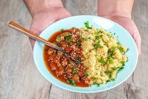Vegetarisches und gesundes Mittagessen: Mann hält einen Teller mit Bohnen in Tomatensoße, gekochtem Reis mit Sesam und Petersilie