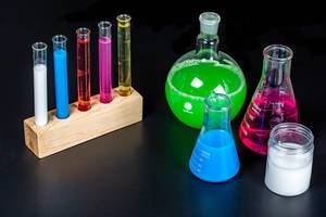 Verschiedene Laborgläser wie Erlenmeyerkolben, Stehkolben und Reagenzgläser mit farbigen Testflüssigkeiten auf schwarzem Hintergrund