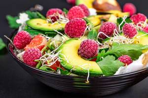 Vitamin Salat mit grünem Salat, Avocado-Scheiben, Himbeeren, Mikrogrün und anderen Zutaten in einer schwarzen Schale Nahaufnahme