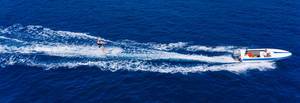 Wassersportler auf Wasserskis, wird von einem Motorsportboot über das blaue Meer gezogen und schlägt weiße Wellen, als Drohnenbild