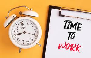 Wecker und ein Klemmbrett mit dem handgeschriebenen Text "Time to work / Zeit zu arbeiten", vor gelbem Hintergrund