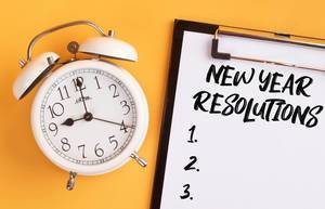 Wecker und ein Klemmbrett mit dem Text "New Year Resolutions / Neujahrsvorsätze" mit einer leeren Liste,  vor gelbem Hintergrund