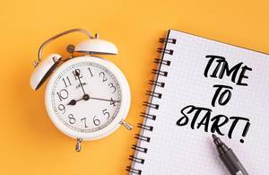 Wecker und ein Notizblock mit dem Text "time to start / Zeit zu beginnen", mit einem Filzstift vor gelbem Hintergrund