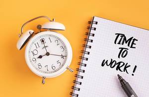 Wecker und ein Notizblock mit dem Text "time to work / Zeit zu arbeiten", mit einem schwarzen Filzstift vor gelbem Hintergrund