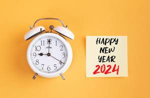 Wecker und ein Zettel mit ‘Happy new year 2024’ Text vor gelbem Hintergrund