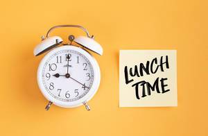 Wecker und ein Zettel mit ‘Lunchtime’ Text vor gelbem Hintergrund