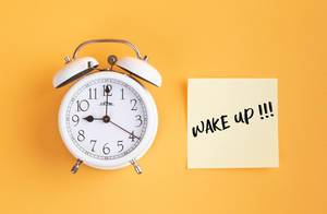 Wecker und ein Zettel mit ‘Wake up!!!’ Text vor gelbem Hintergrund