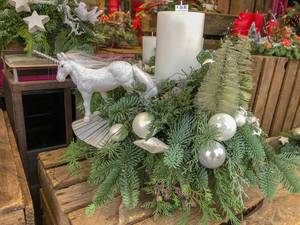 Weihnachtsgesteck mit Tannenzweigen, weißen Kerzen, perlmuttfarbenen Weihnachtskugeln und silbernem Einhorn in Verkaufsauslage, im Hintergrund weitere Weihnachtsdekoration und Adventskränze mit Sternen und Kerzen in rot