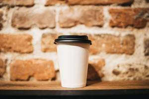 Weißer Coffee-to-go Kaffeebecher aus Papier mit schwrzem Deckel vor Backsteinwand