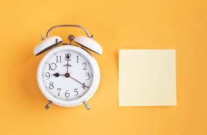 White alarm clock on yellow background with empty sticky note