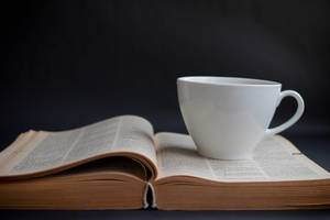 White Ceramic Coffee Cup on Old Open Book on Black Background