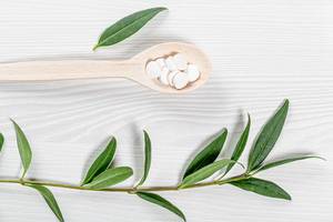 White tablets in a wooden spoon and a branch with green leaves on a white wooden background