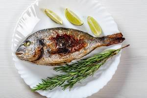Whole baked Dorado fish on a white plate with fresh rosemary and lime slices