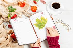 Woman puts maple leaf in empty notebook on autumn background of white table with scarf, coffee Cup and glasses