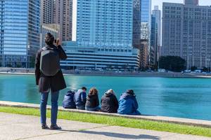 Woman uses her smartphone to take a photo of the Chicago skyscrapers by the shoreline while others sit by the water