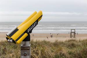 Yellow-black binoculars with inscription Galaxy in front of the beach of Zandvoort with giant chair