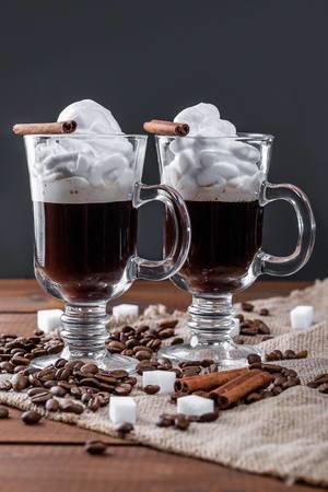 Zwei Kaffee im Glas, mit geschlagener Sahne, Zuckerwürfeln, Zimtstangen und Kaffeebohnen, auf einem Holztisch