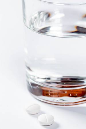 Zwei Tabletten vor gefülltem Wasserglas vor weißem Hintergrund